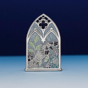 May include: A silver-toned metal enamel pin shaped like a pointed gothic arch window. The design features a moth with patterned wings, green leaves, and blue flowers set against a swirling light gray background with dark glitter accents.