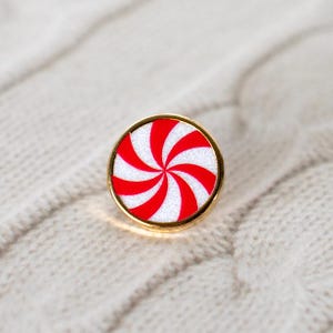 May include: A circular pin with a gold border, showcasing a red and white peppermint candy design. The pin has a spiral pattern of red stripes on a white base. The background is a textured, off-white fabric.