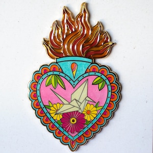 Peut inclure: Épingle en émail en forme de cœur avec un contour doré et un motif de flamme en haut. L'épingle présente un cœur rose avec une grue en origami blanche, des fleurs jaunes et roses et des feuilles vertes. La bordure est turquoise et orange.