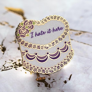 May include: A decorative enamel pin in the shape of a cake with gold trim and purple accents. The pin features the phrase "I hate it here" in purple script. The top of the cake is decorated with purple roses.