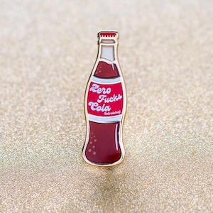 Könnte beinhalten: Goldfarbene Emaille-Anstecknadel in Form einer Sodaflasche mit dem Text "Zero Fucks Cola" in Weiß auf rotem Hintergrund. Der Text "Refreshing!" steht in kleinerer weißer Schrift unter der Cola.
