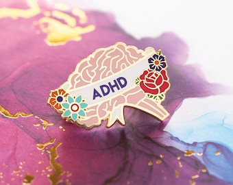 May include: A pink enamel pin with a brain shape and the word "ADHD" on a white banner. The brain is decorated with colorful flowers.