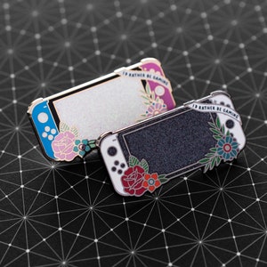 May include: Two enamel pins shaped like the Nintendo Switch console. The top pin is white with blue accents and glitter, and the bottom pin is black with silver accents and glitter. Both pins have floral designs and the text "I'd rather be gaming".