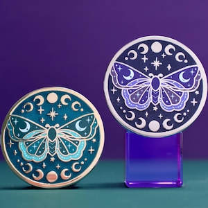 May include: Two circular enamel pins featuring a moth design with celestial motifs. One pin is teal with light blue accents, while the other is dark blue with purple accents. Both pins display a central moth surrounded by moon phases and stars.