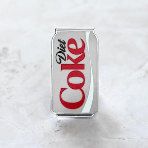 May include: A Diet Coke can-shaped pin with a silver border. The can is light grey with the words "Diet" and "Coke" in red script. A white curved stripe runs down the right side of the can.