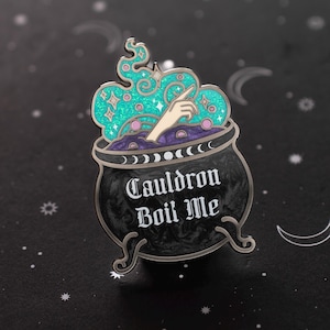 May include: Enamel pin featuring a black cauldron with the text "Cauldron Boil Me" in white. The cauldron has a purple interior with a hand reaching out of it. Above the cauldron is a teal cloud with stars and geometric shapes.
