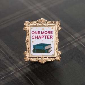 May include: A gold-colored pin with an ornate frame design. The pin features the text "ONE MORE CHAPTER" in burgundy above a stack of two books. The books are teal and green, with gold star accents.