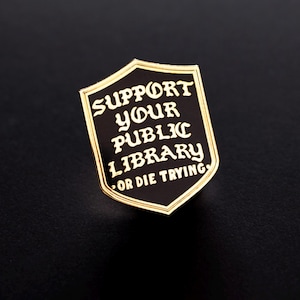 以下が含まれることがあります： 黒と金のシールド型のエナメルピン。ピンには、白い文字で「SUPPORT YOUR PUBLIC LIBRARY OR DIE TRYING」と書かれています。ピンは金の縁取りがあり、黒い背景に配置されています。