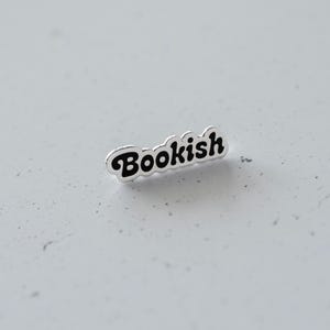 Puede incluir: Un pin de plata con la palabra "Bookish" en letras negras.