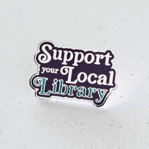 May include: A purple enamel pin with white lettering that reads "Support your Local Library" in a retro font. The word "Library" is in teal.