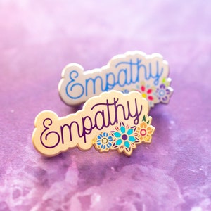 May include: Two enamel pins with the word "Empathy" in a cursive font. The pins are off-white with a floral design in shades of blue, orange, and purple. One pin has blue lettering, and the other has purple lettering.