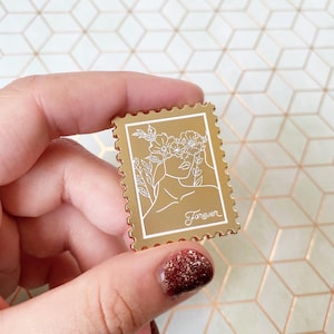 May include: A gold-coloured enamel pin in the shape of a postage stamp. The pin features a white line drawing of a woman's face with a floral crown and the word "Forever". The pin has a scalloped edge.