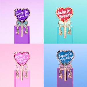 May include: Four heart-shaped enamel pins shaped like lollipops with gold-toned metal outlines. Each pin features a bow at the base and white text: Sucker for Fictional Men, Sucker for Yearning, Sucker for Friends to Lovers, and Sucker for Fake Dating.