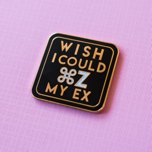 May include: A square black enamel pin with gold trim and lettering. The text reads "WISH I COULD ⌘Z MY EX" in gold. The "⌘Z" symbol is silver. The pin is set against a pink background.