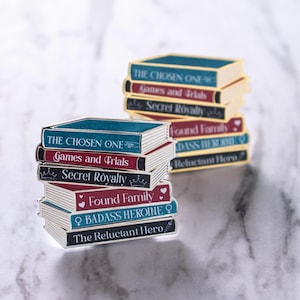 May include: Stacks of enamel pins shaped like miniature books. Each pin features a different title, including "The Chosen One," "Games and Trials," and "Secret Royalty." The pins have gold edges, teal and burgundy covers, and white pages, creating a detailed, miniature book appearance.