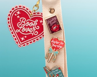 May include: A cream-colored fabric wristlet keychain featuring a gold ring and a red heart-shaped embroidered patch that says Book Lover. Three enamel pins are attached to the strap: a book labeled Reading Smut, a heart lollipop labeled Sucker for Yearning, and a book held by a hand labeled Do not Disturb I'm Busy Reading.