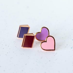 Puede incluir: Tres pines de esmalte con un libro morado, un libro marrón y un corazón rosa. Los pines tienen un contorno dorado.