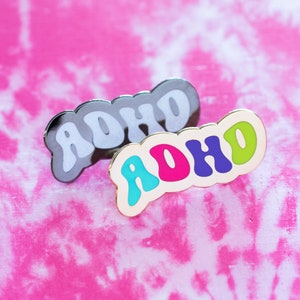 May include: Two pins with the word "ADHD" written in a colourful, bubbly font. The top pin is black with white letters, and the bottom pin is gold with turquoise, pink, purple, and green letters.