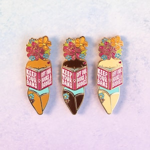 May include: Three enamel pins featuring stylized figures with floral headdresses and holding books. The books read "KEEP YOUR BOOKS & BODIES OFF OUR BANS." The figures are in shades of tan, brown, and cream, wearing blue underwear.