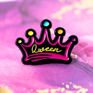 May include: A black enamel pin shaped like a crown with pink and yellow accents. The word "Queen" is written in yellow inside the crown.