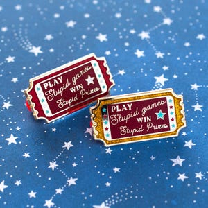 Puede incluir: Dos pines de esmalte con forma de boletos vintage. Los pines son rojos y dorados con el texto "PLAY Stupid games WIN Stupid Prizes". El fondo es azul oscuro con estrellas blancas.