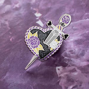 Puede incluir: Un pin de esmalte plateado con un corazón negro atravesado por una daga plateada. El corazón tiene un diseño floral morado y amarillo.