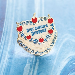 May include: A gold-edged enamel pin in the shape of a cake, decorated with red cherries and blue accents. The pin features the words "Diet Culture Dropout" in blue text on a cream-colored background.