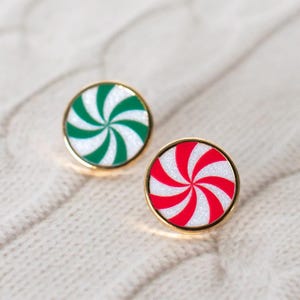 May include: Two round, gold-rimmed pins with a peppermint candy design. One pin features green and white stripes, while the other has red and white stripes. The pins are set against a textured, off-white background.