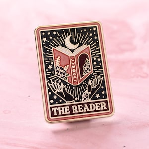 May include: A rectangular enamel pin with a tarot card design. The pin features a black background with a crescent moon, stars, and an open book. The book has floral designs and is held by two hands. The text "THE READER" is at the bottom.