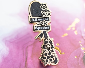 May include: A gold-coloured metal pin with black enamel and the text "All out of Spoons". The pin features a heart-shaped top with glitter, floral accents, and a ribbon design. The pin is on a pink and gold surface.