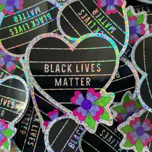 May include: Holographic stickers with a heart shape and the text "BLACK LIVES MATTER". The heart is black with horizontal lines and a silver glitter border. Each sticker has a flower with purple and pink petals.