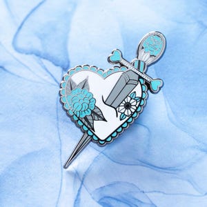 May include: A silver enamel pin featuring a white heart with a blue flower and a silver dagger piercing through it. The dagger has a blue rose on the handle.