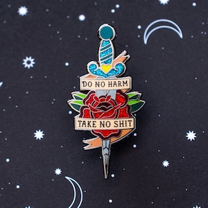 May include: Enamel pin showing a dagger through a red rose. The pin has a teal handle with glitter and a yellow heart. The banner reads "DO NO HARM" and "TAKE NO SHIT" on a black background with stars and crescent moons.