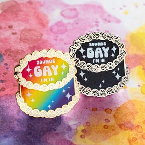 Könnte beinhalten: Zwei Kuchenförmige Emaille-Anstecknadeln mit goldenen Rändern. Einer ist regenbogenfarben mit dem Text "SOUNDS GAY I'M IN". Der andere ist schwarz mit dem gleichen Text in Weiß. Beide haben Sternakzente.