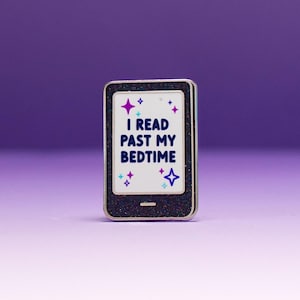 Könnte beinhalten: Eine rechteckige Emaille-Anstecknadel in Form eines E-Readers mit schwarzem Glitzerrahmen und weißem Bildschirm. Auf dem Bildschirm steht der dunkelblaue Text "I READ PAST MY BEDTIME", umgeben von kleinen violetten und hellblauen Sternsymbolen.