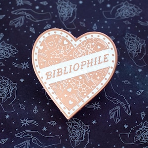 May include: Rose gold heart-shaped pin with a white outline and the word "Bibliophile" written across the center. The pin is decorated with a floral pattern.