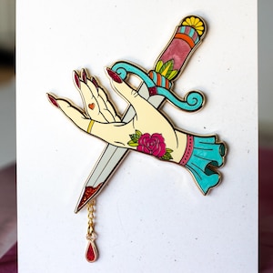 May include: A decorative pin featuring a hand with red fingernails, a gold ring, and a heart tattoo, pierced by a dagger. The dagger has a teal handle and a red, glittery drop. A rose tattoo is on the hand.