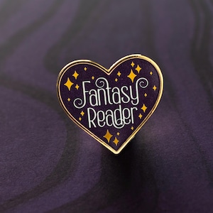 以下が含まれることがあります： 金色の縁取りが付いたハート型のエナメルピン。「Fantasy Reader」の文字が白い筆記体で、濃い紫色の背景に黄色の星のアクセントで描かれています。このピンは、おそらく収集品またはアクセサリーです。