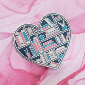 May include: A heart-shaped enamel pin with a bookshelf design. The bookshelf is filled with books, plants, and other small objects. The text "Read Queer Stories" is printed on the pin.