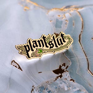 May include: A gold-colored enamel pin with the text "plantslut" in a stylized font. The pin features green leaves and a red and green flower design. The pin is set against a light blue and gold marbled background.