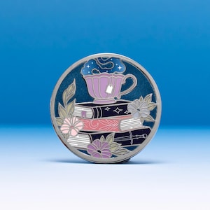 May include: A circular enamel pin featuring a stack of three books topped with a light purple teacup containing a swirling dark blue starry liquid. The design includes light pink and purple flowers, green leaves, and a silver sword outline.