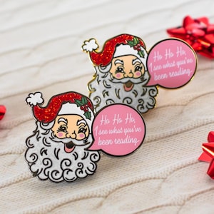 Puede incluir: Dos pines de esmalte de Papá Noel con sombreros rojos con purpurina y bocadillos. Los bocadillos rosas dicen "Ho Ho Ho, I see what you've been reading". Los pines tienen barbas blancas y están sobre una superficie de punto blanca.