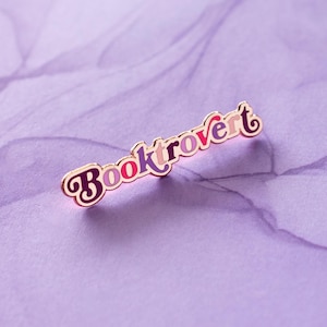 May include: A gold-colored enamel pin with the word "Booktrovert" in colorful letters. The letters are in a retro font style.