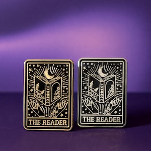May include: Two rectangular enamel pins featuring a black background with gold and silver metallic line art. The design depicts an open book, a crescent moon, stars, and hands, with the text THE READER printed at the bottom of each pin.