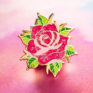 Puede incluir: Un broche de esmalte con forma de rosa. El broche presenta una rosa rosa brillante con pétalos rosa claro y hojas verdes con ribetes dorados. El diseño de la rosa es detallado, perfecto para añadir un toque floral a cualquier atuendo o accesorio.