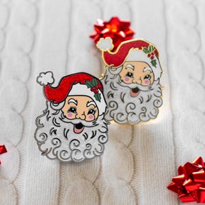 May include: Two Santa Claus enamel pins with red hats and white beards. The pins feature a smiling Santa face with holly accents on the hat. The pins are set against a white, textured background with red bows.