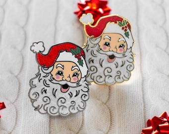 May include: Two Santa Claus enamel pins with red hats and white beards. The pins feature a smiling Santa face with holly accents on the hat. The pins are set against a white, textured background with red bows.