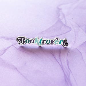 May include: A silver enamel pin with the text "Booktrovert" in a colorful, retro font. The letters are outlined in white and filled with shades of purple, blue, and green.
