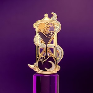 May include: A gold-toned metal enamel pin shaped like an hourglass. The design features a textured gold sun at the top and a white crescent moon at the bottom, set against a deep purple background with small star accents and swirling white details.