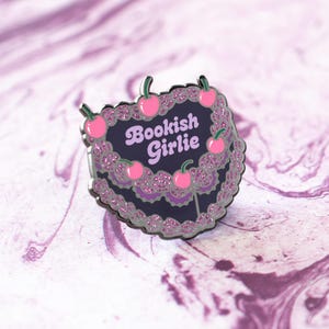 Könnte beinhalten: Eine schwarze und rosafarbene Emaille-Anstecknadel in Form eines Kuchens mit dem Text "Bookish Girlie" und rosa Kirschen am Rand.